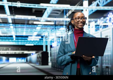 Smiling data center female engineer configuring server settings with laptop software. Happy woman in modern high tech server hub using notebook to do maintenance tasks on infrastructure machines Stock Photo
