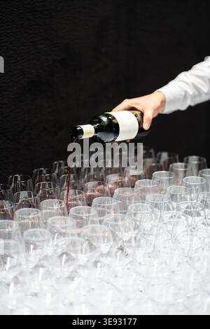 Hand pouring red wine from a bottle into rows of empty glasses, preparing for a large event or wine tasting session Stock Photo