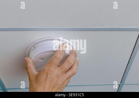 hand installing or checking smoke detector on ceiling tile indoors. Fire safety maintenance, home protection and alarm system concept. Stock Photo