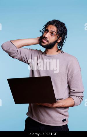 Arab programmer holding laptop, uncertain about software app development. Young person rubbing head and looking away while thinking about business report on portable computer Stock Photo