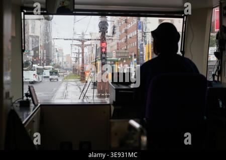 A moody POV from inside a Kagoshima tram on a rainy day. The driver's dark silhouette and rain-streaked glass highlight a nostalgic urban journey. Stock Photo