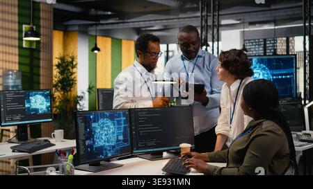 Multiethnic technicians use devices in AI startup office to train predictive models for language processing tasks. Multiracial coworkers use deep learning technical expertise for tasks, camera A Stock Photo