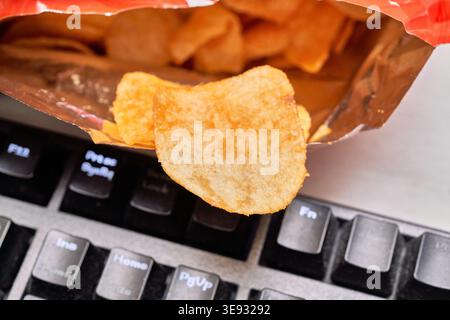A close-up photo of a crispy potato chip over a black computer keyboard. This image perfectly captures the relatable habit of snacking at the desk. Stock Photo