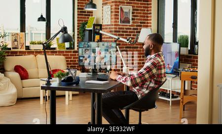 Cheerful employee in video conference meeting chatting with diverse international team while working from home. Man teleworking in online video call with coworkers remotely from cozy apartment Stock Photo