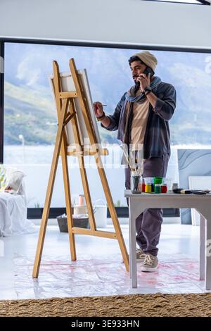 Adult male painter standing at easel in studio, wearing beanie and scarf, holding brush and phone Stock Photo