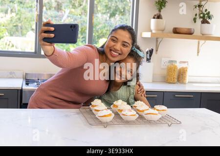Indian mother and child posing smartphone selfie in kitchen by frosted cupcakes, wearing blue bow Stock Photo