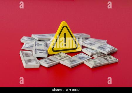 A yellow warning triangle and stacks of US dollar bills on a red background. Financial risk, economic danger, inflation, or investment alerts concept. Stock Photo