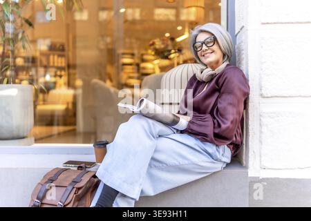 Smiling mature woman with grey hair and eyeglasses sitting on a city ledge and reading a magazine, enjoying a relaxed moment with coffee and headphone Stock Photo