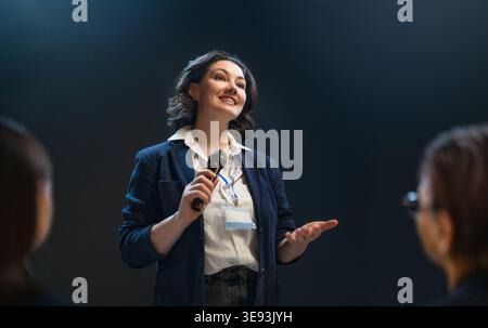 Dynamic image of a confident businesswoman giving a public speech at a professional event. She stands with poise and smiles, holding a microphone and Stock Photo