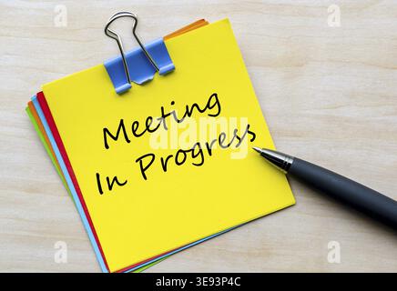 Meeting In progress handwritten text on yellow notepad with silver clip placed on wooden desk Stock Photo