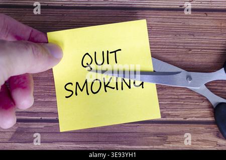 Hand holding a yellow sticky note with the words Quit Smoking written on it, while a pair of scissors is cutting through the note Stock Photo
