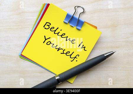 Believe In Yourself handwritten text on yellow notepad top view with silver clip on wooden desk Stock Photo