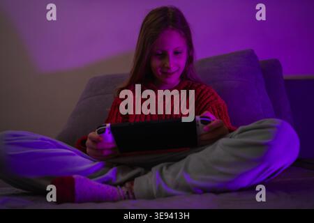 Smiling girl playing a video game on a portable device illuminated by purple ambient light Stock Photo
