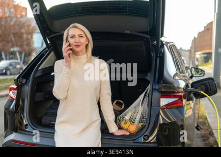 A woman talks on her smartphone next to the trunk of an electric car, which is plugged into a charging station. A mesh bag filled with fresh produce is visible inside the trunk. Stock Photo