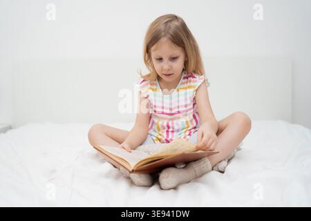 A smiling girl sits cross-legged on a bed reading a large book with both hands in a bright minimalist room cozy home learning childhood literacy Stock Photo