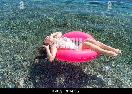 Summer vacation relaxation young woman in white swimsuit floating on pink inflatable ring in clear sea water on sunny day Stock Photo