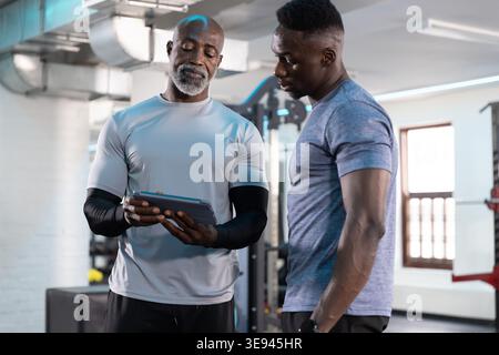 African American male trainer and client wearing gray shirts reviewing tablet in gym with plates Stock Photo