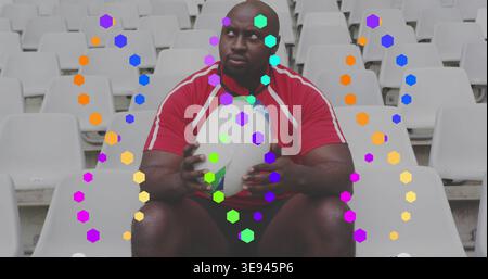 Sitting athlete wearing red jersey holding white rugby ball among stadium white seats with hexagons Stock Photo