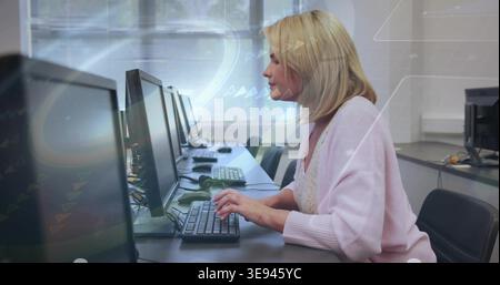 Typing mid-adult woman wearing pink cardigan at lab with monitors and keyboards, blinds, copy space Stock Photo