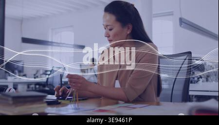 Working Korean designer in tan blouse using stylus on tablet at open office, holding color swatches Stock Photo