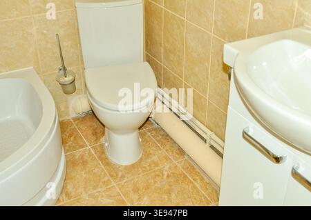 A clean and functional bathroom featuring a white toilet, a vanity sink, and a bathtub, all set against beige tiled walls and floor Stock Photo