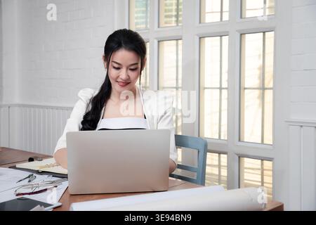 Charming smiling motivated woman architect working on building plan using laptop computer. Engineer designing virtual sketch for futuristic constructi Stock Photo
