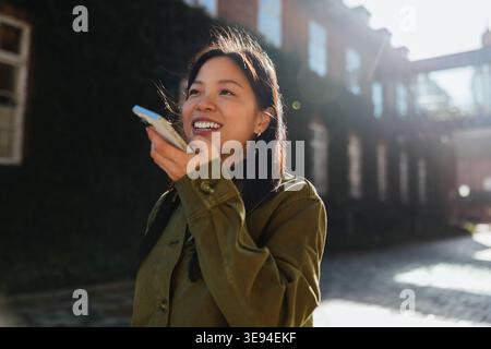 Young woman speaks into her phone while walking outdoors on a sunlit city street, smiling and holding the smartphone close to her mouth as brick build Stock Photo
