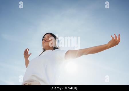 Woman standing outdoors with eyes closed and arms outstretched, wearing headphones and a white t-shirt against a bright blue sky, basking in sunlight Stock Photo
