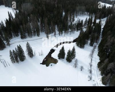 Aerial view of the Dolomites featuring a winding dark stream, dense evergreen forests and people on a small bridge in the snow, Rocca Pietore, Veneto, Stock Photo