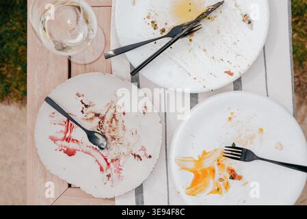 Empty plates show messy meal leftovers. Forks and spoons rest beside smudged, colorful stains. Glass holds faintly cloudy beverage. Wooden table holds Stock Photo
