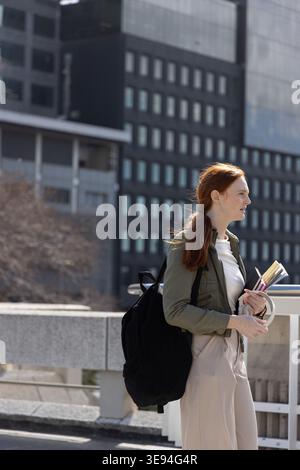 Woman standing on bridge in city holding notebooks and pen, carrying black backpack, copy space Stock Photo