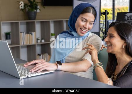 Two women colleagues one wearing blue hijab working on laptop at meeting table with smartwatch Stock Photo