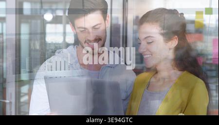 Working coworkers wearing mustard blazer, hoodie viewing tablet in office by glass with postits Stock Photo