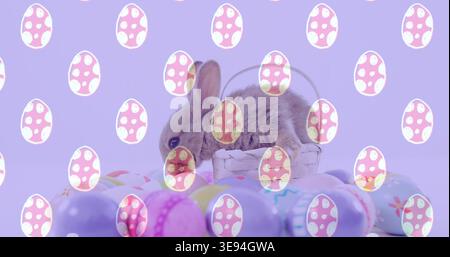 Sitting small brown rabbit on tabletop among whitewashed wicker basket, pastel eggs and egg-overlay Stock Photo