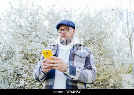 Overweight bearded Caucasian man using smartphone near blooming tree in spring Stock Photo