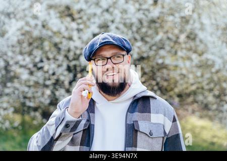 Happy overweight bearded Caucasian man talking on smartphone near blooming tree Stock Photo