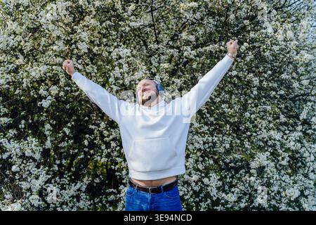 Joyful bearded man wearing headphones raising arms near blooming tree Stock Photo