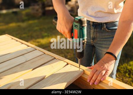 Young woman using a screwdriver while working in a workshop outdoors. Female worker using drill on wooden plank while working at construction site Stock Photo