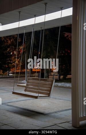 Wooden swing bench hanging on chains in a modern urban space at night, illuminated by soft warm lighting. Stock Photo
