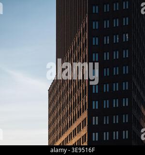 Dark brick skyscraper facade against blue sky. Tall dark brick building with a repetitive window pattern, partially lit by warm setting sun. Stock Photo