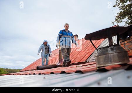 Two men repairs chimney on house roof. Roofer working on metal rooftop construction for home maintenance. Professional worker repairing ventilation sy Stock Photo