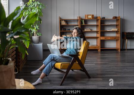 Happy satisfied with work done woman stretching hands, relaxing in cozy armchair with laptop in home office. Smiling girl resting after finished remote freelancer tasks on computer dreaming take break Stock Photo