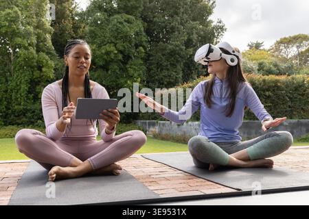 African American instructor guiding Asian woman on brick patio on yoga mats using tablet and VR. Backyard, outdoor, balance, fitness, wellness, training, mindfulness Stock Photo