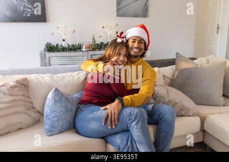 Couple hugging while sitting on sectional sofa at home wearing Santa hat and reindeer headband. Cozy, festive, holiday, romantic, intimate, warm, soft Stock Photo