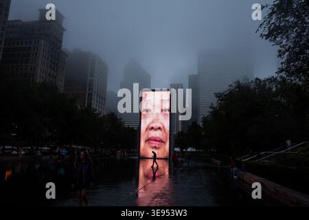 A person walks through a wet plaza at night, with a large digital billboard displaying a face in the background. Stock Photo