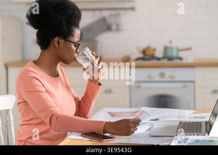 Young african businesswoman work from home, drink water using laptop computer in remote office in kitchen. Female freelancer, designer or architect planning project blueprint remotely during covid-19 Stock Photo