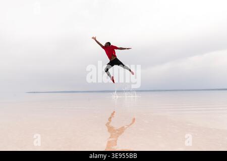 Energetic young man captured mid-air while jumping across a reflective salt flat landscape. Dynamic action scene representing freedom, joy, movement, Stock Photo