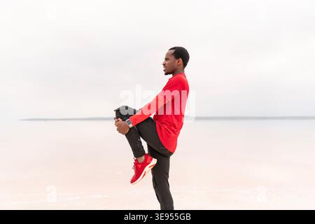 Full-body image of a young man stretching his leg before or after exercise in a minimalist outdoor setting. Fitness, flexibility, warm-up routine, and Stock Photo