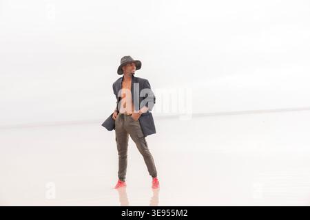 Fashion portrait of a stylish young man wearing an open blazer and hat while standing in a minimalist outdoor setting. Modern fashion, confidence, ind Stock Photo