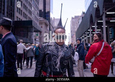 A person with a large mohawk and sunglasses walks down a busy city street while wearing a studded leather jacket. Stock Photo